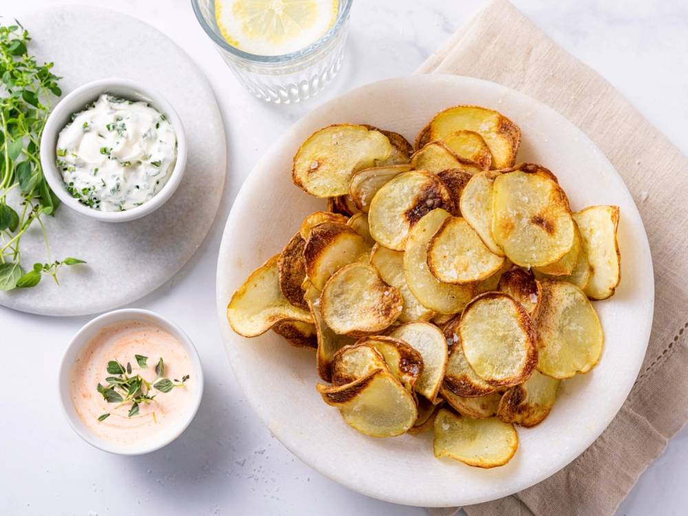 Sprøstekte potetgull laget i airfryer på en hvit tallerken med to små dippsauser og en glass med vann med sitron, på et lyst bord.