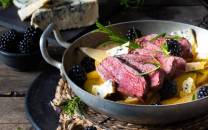 Stekt kjøtt med bær og sitron i en hvit gryte, omgitt av frukt og urter på et bord.