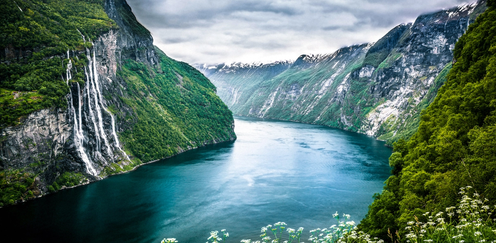 En fjord med høye, grønne fjell på begge sider og fossefall som renner ned i vannet.