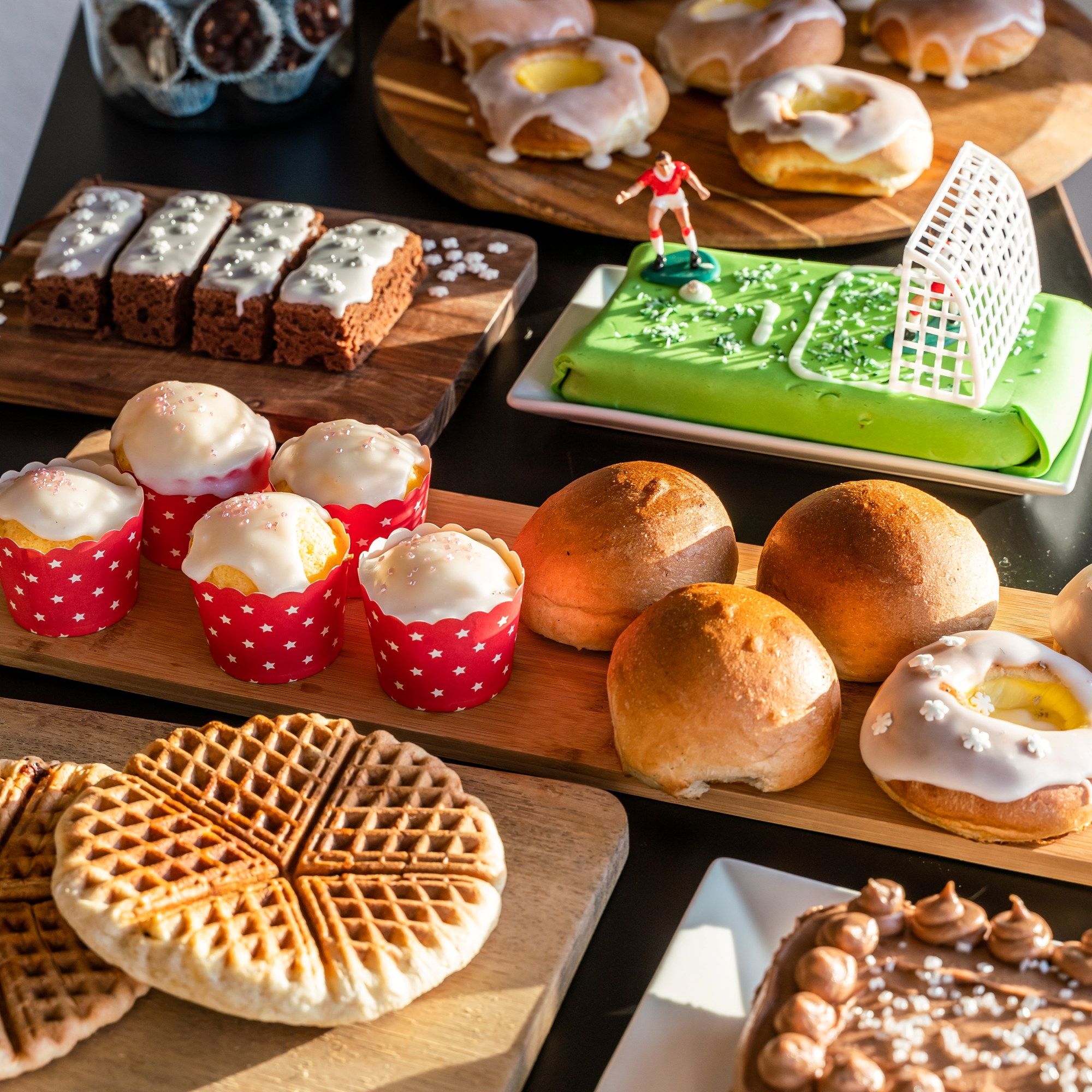 Et utvalg av ulike kaker og bakverk på et bord, inkludert cupcakes, vafler, boller og en marsipankake med fotballtema.