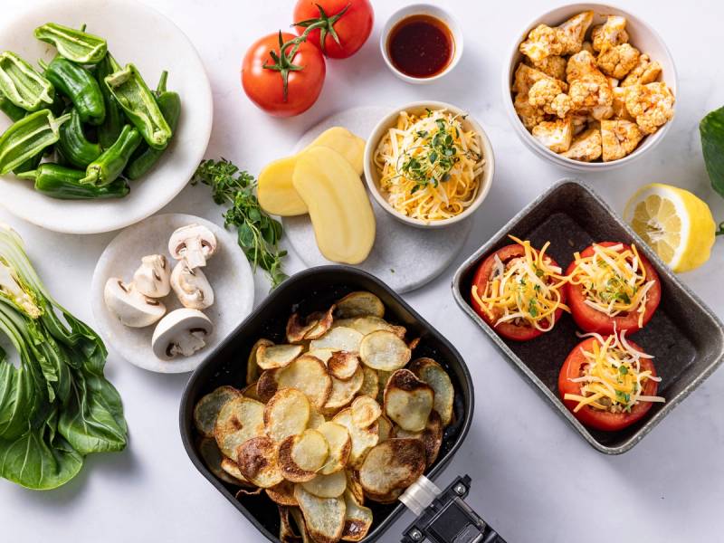 Ulike matretter og ingredienser som poteter, tomater, sopp, grønne blader, og ferdiglaget mat i en airfryer, sannsynligvis tilberedt med en airfryer.