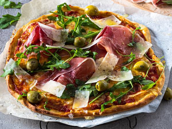En pizza med skinke, grønne oliven, ruccola og ost på en bakepapir, klar til servering.