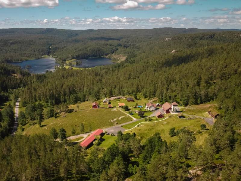 En oversikt over en liten landsby med spredte hus omgitt av grønne skoger og vann i bakgrunnen.