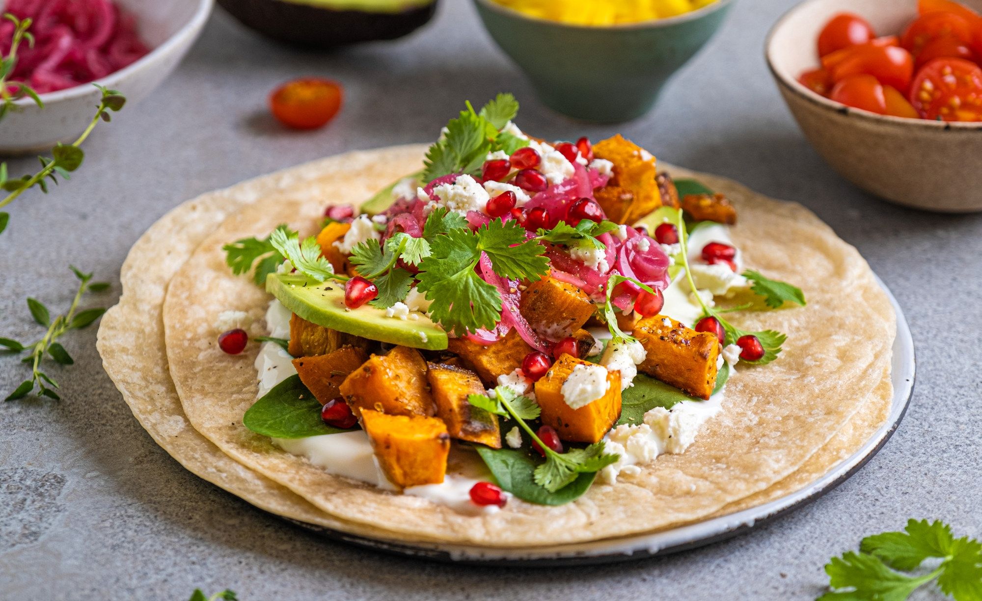Vegetarisk taco med søtpotet, avokado, koriander og granateple på en tortilla, omgitt av små boller med cherrytomater og andre grønnsaker