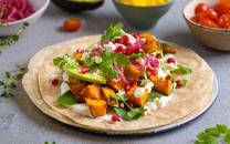 Vegetarisk taco med søtpotet, avokado, koriander og granateple på en tortilla, omgitt av små boller med cherrytomater og andre grønnsaker
