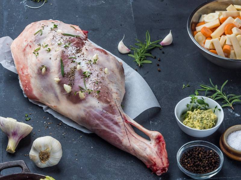 Et stykke lammelår med krydder og urter, klar til å bli tilberedt, omgitt av ulike krydder og grønnsaker på en mørk overflate.