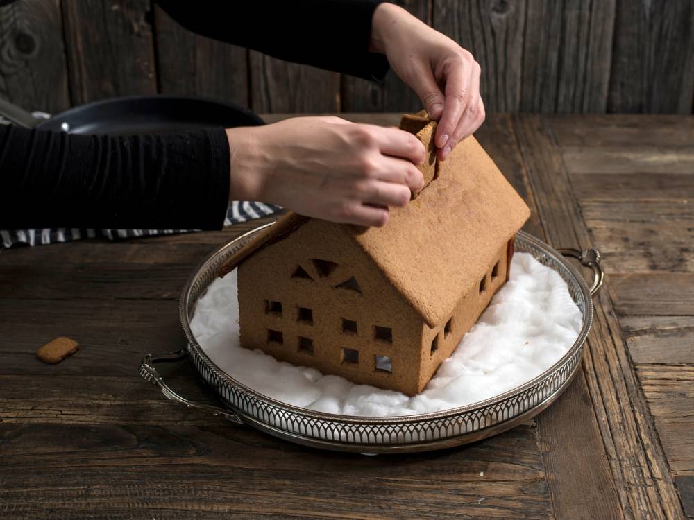 Hender dekorerer et pepperkakehus med små vinduer og dører på et fat med mel, foran en trevegg.