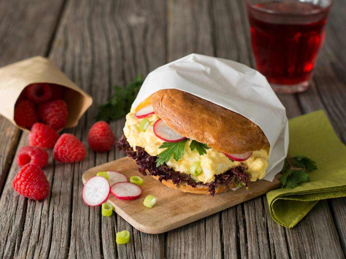 Rundstykke med eggsalat, skiver av radise og grønt salat på en skjærebrett, med et glass rød drikke i bakgrunnen.