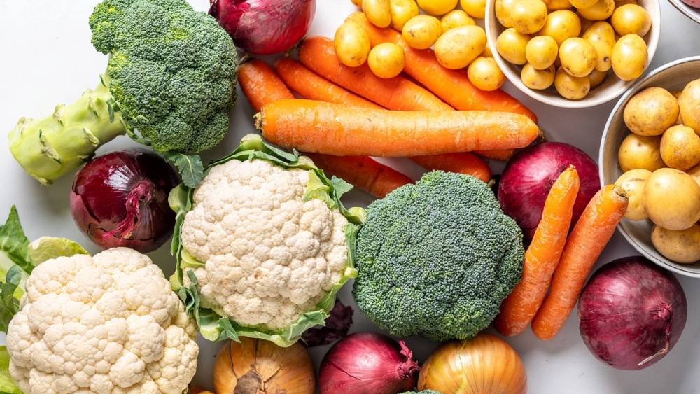 Et utvalg av ulike ferske grønnsaker, inkludert brokkoli, blomkål, gulrøtter, rødløk og kikerter, på en hvit bakgrunn.