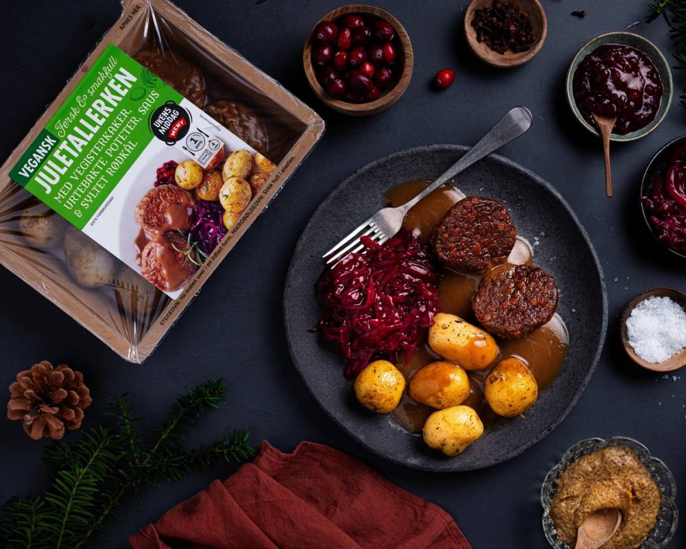 En vegansk julemiddag med kjøttfrie kjøttboller, rødkål, poteter og lingonsyltetøy på en svart tallerken, med en pakke vegansk julemat i bakgrunnen.