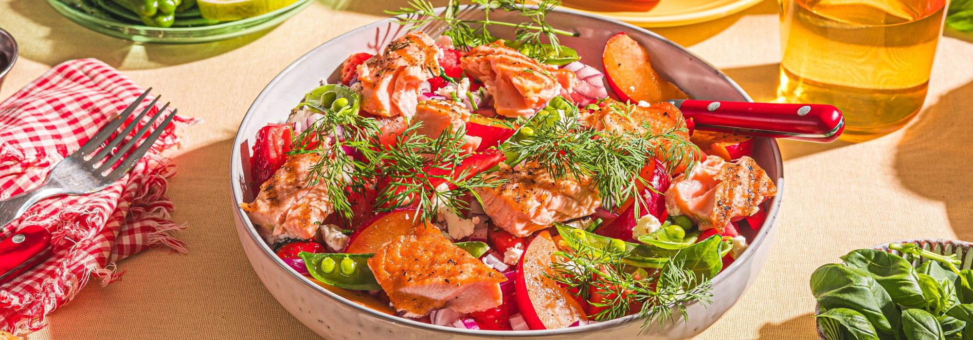 En bolle med en salat av tomater, agurk, urter og sitronbåter, omgitt av glass med drikke og bestikk på et bord.