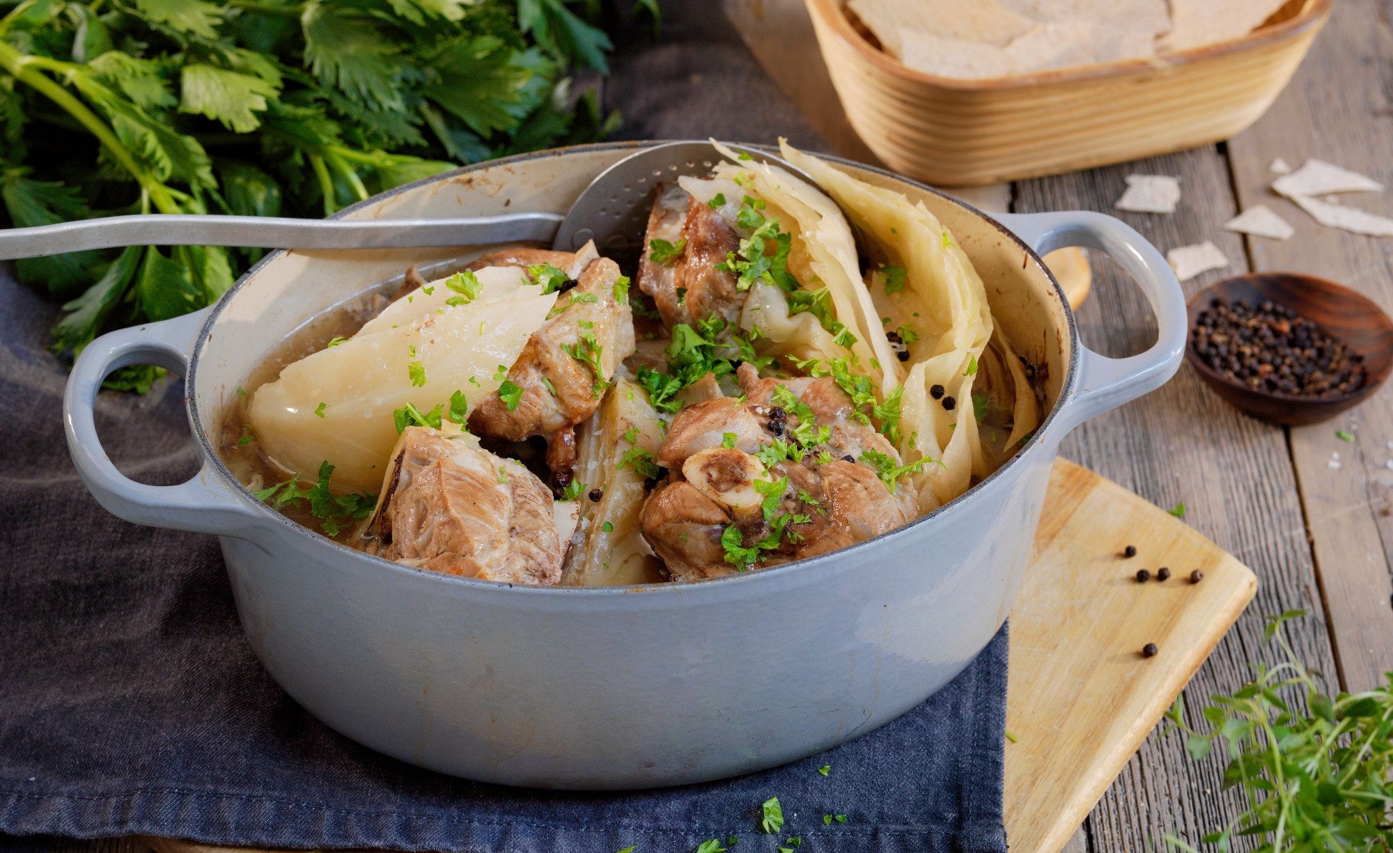 Fårikål i en hvit gryte med hel kål og kjøtt, pyntet med hakket grønt, på et bord med urter og krydder rundt.