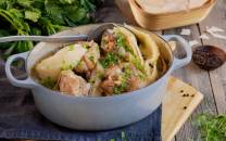 Fårikål i en hvit gryte med hel kål og kjøtt, pyntet med hakket grønt, på et bord med urter og krydder rundt.