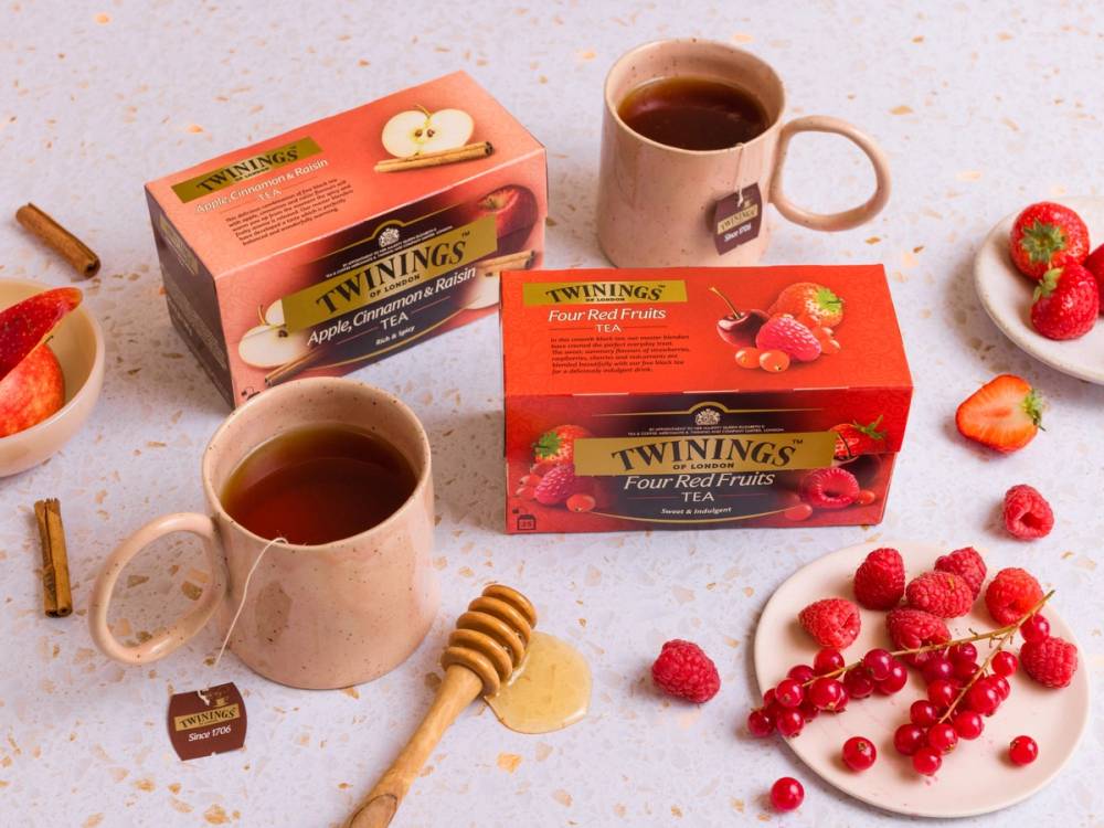 To kopper med te, to Twinings te-esker med bærsmak, og friske jordbær og bringebær på et bord.