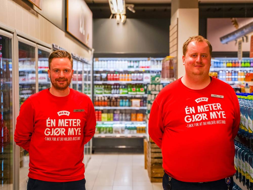To personer i røde t-skjorter står i en dagligvarebutikk med hyller fylt med drikkevarer i bakgrunnen.