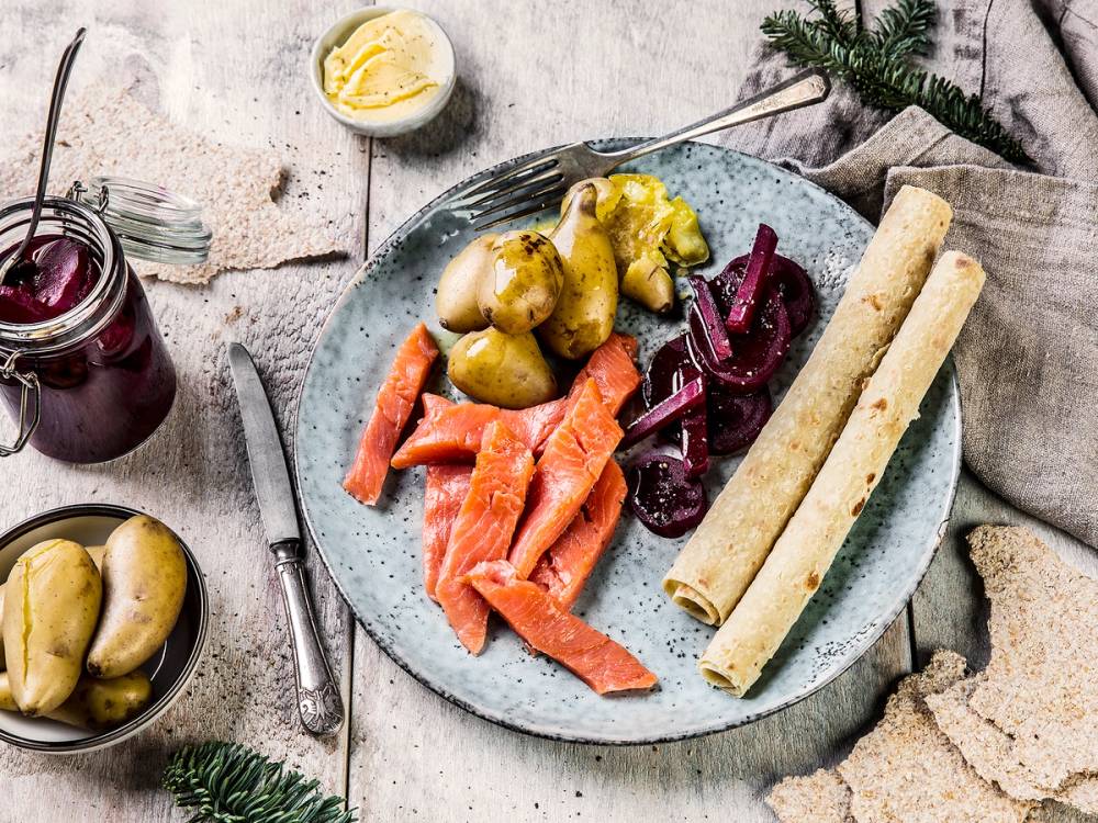 En tallerken med rakfisk, lefser, syltede grønnsaker og rødbeter, omgitt av små glass med tilbehør, på et bord med juledekorasjoner.