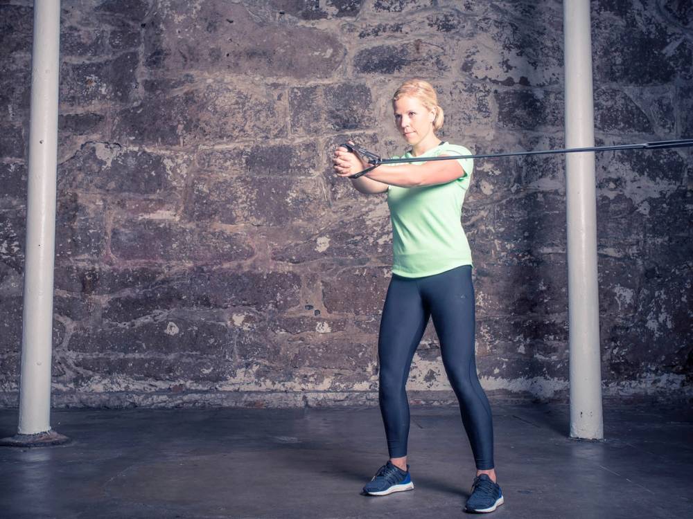 En person utfører en pallof press-øvelse med strikk foran en murvegg, iført treningstøy.