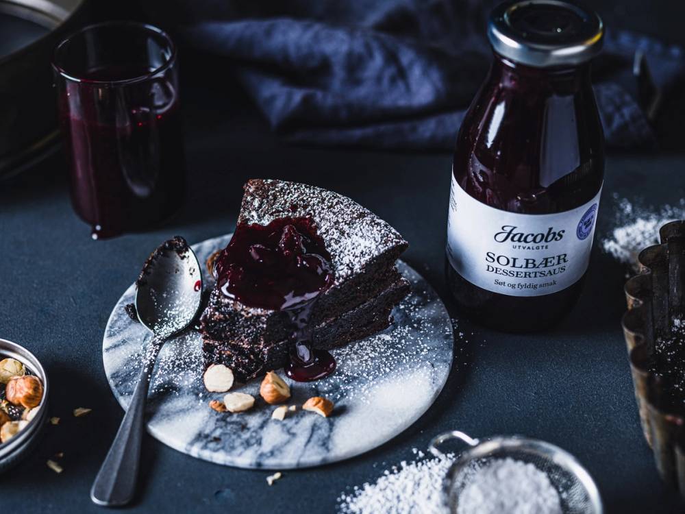Et stykke sjokoladekake med rød saus og et glass med mørk drikke på et mørkt bord, med en Jacobs Solberg-flaske ved siden av.