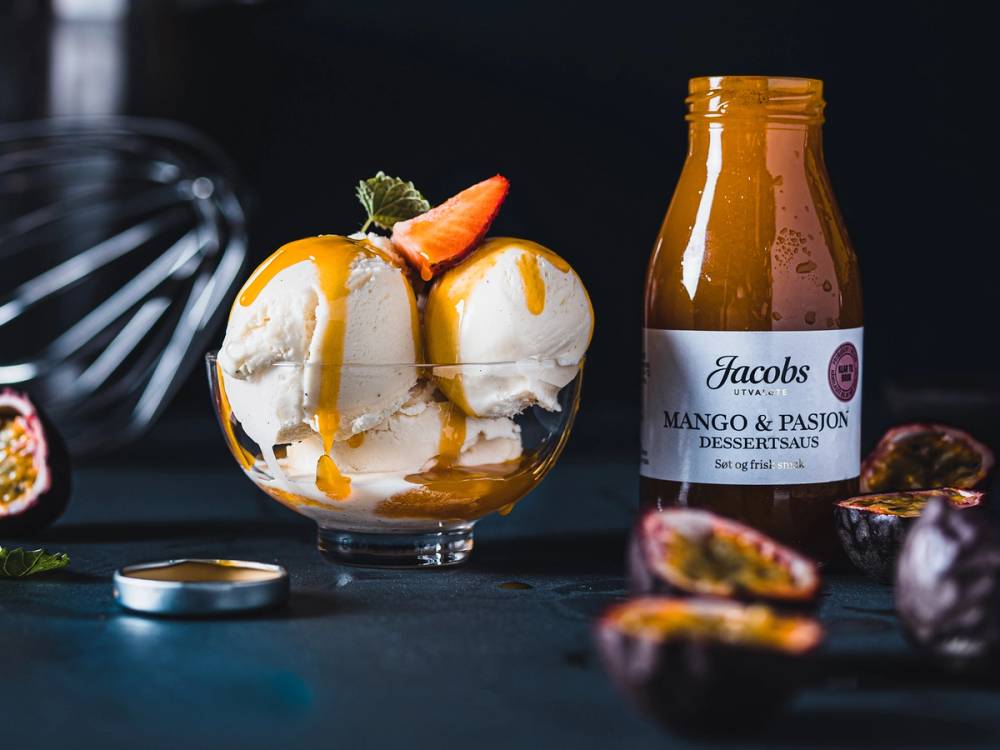Is med mango og pasjonsfrukt i en glassbolle med en flaske Jacobs mango og pasjonsfrukt-dressing ved siden av, omgitt av pasjonsfrukt og en skje.