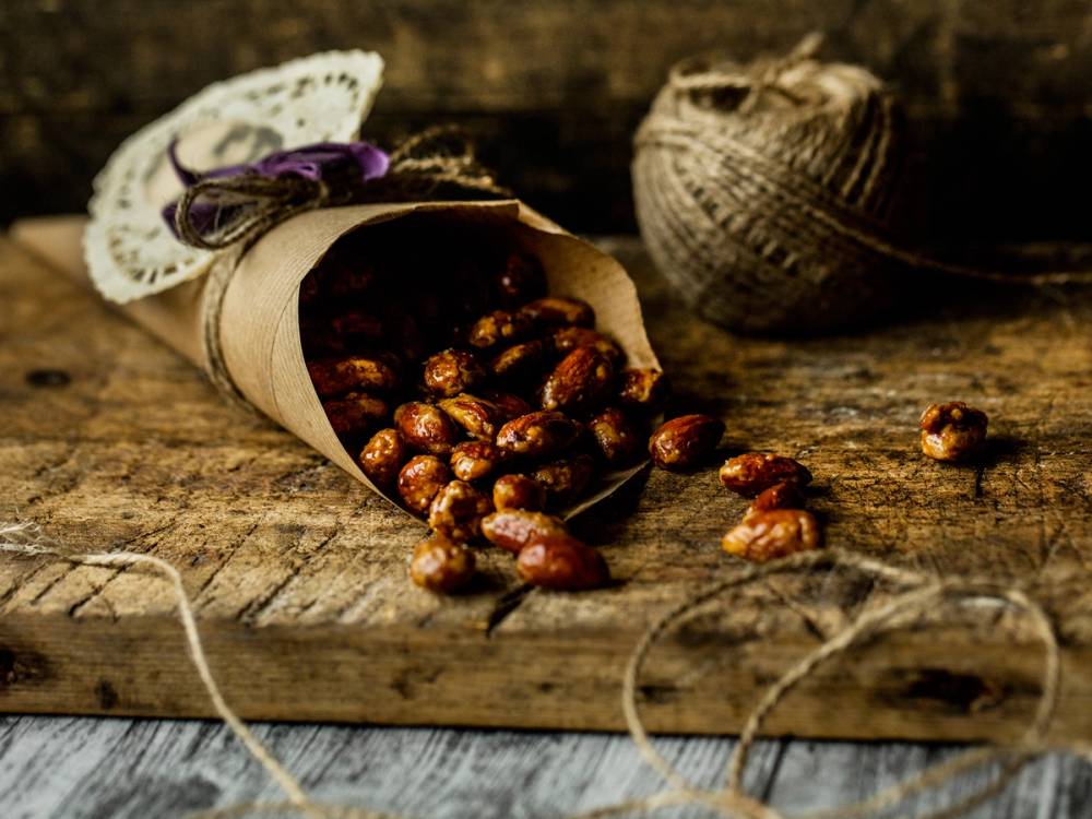 Brente mandler i en papirpose på et trebord med en keramikkrukke i bakgrunnen.