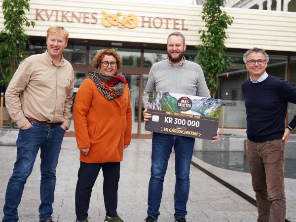 Fire personer står foran Kvikknes Hotel, hvor en av dem holder et stort sjekk med teksten 'KR 300 000' og 'Matskattstipendet 2020'.