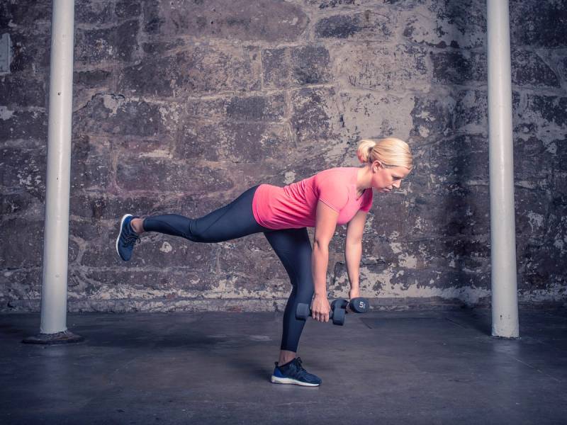 En kvinne utfører en ettbeins strakmark med en manual i hånden foran en murvegg.