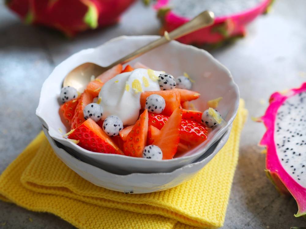 Skål med marinerte jordbær og en klatt kesam, med en liten skje oppi, plassert på en gul duk.