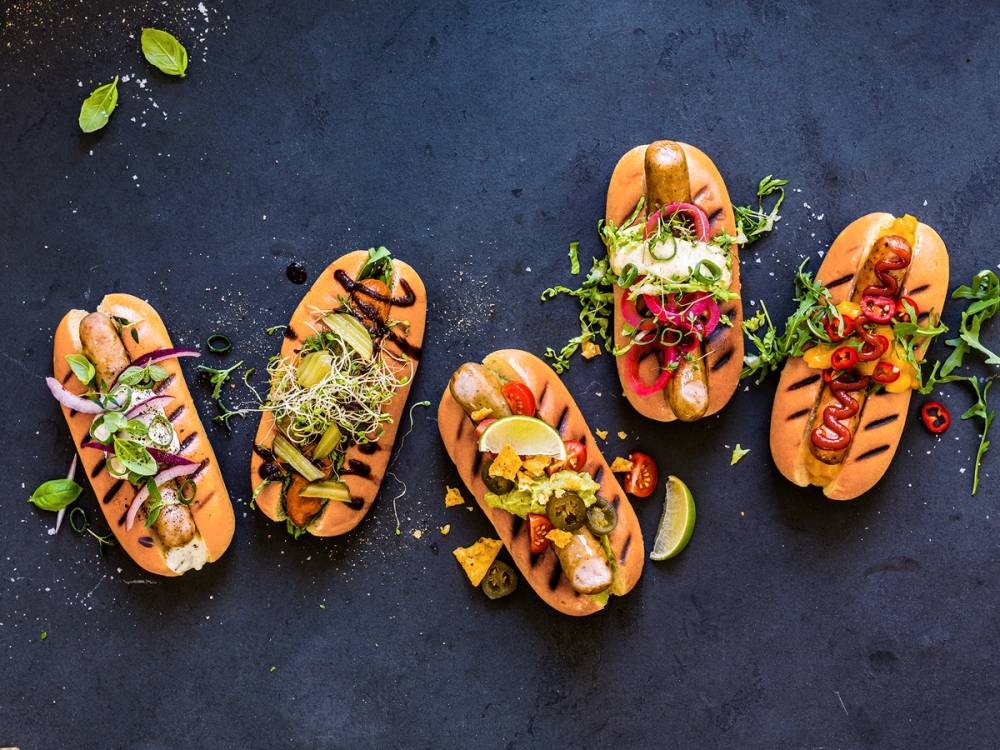 Fem ulike typer grillpølser med tilbehør som løk, chili, sitron og urter på en mørk overflate.