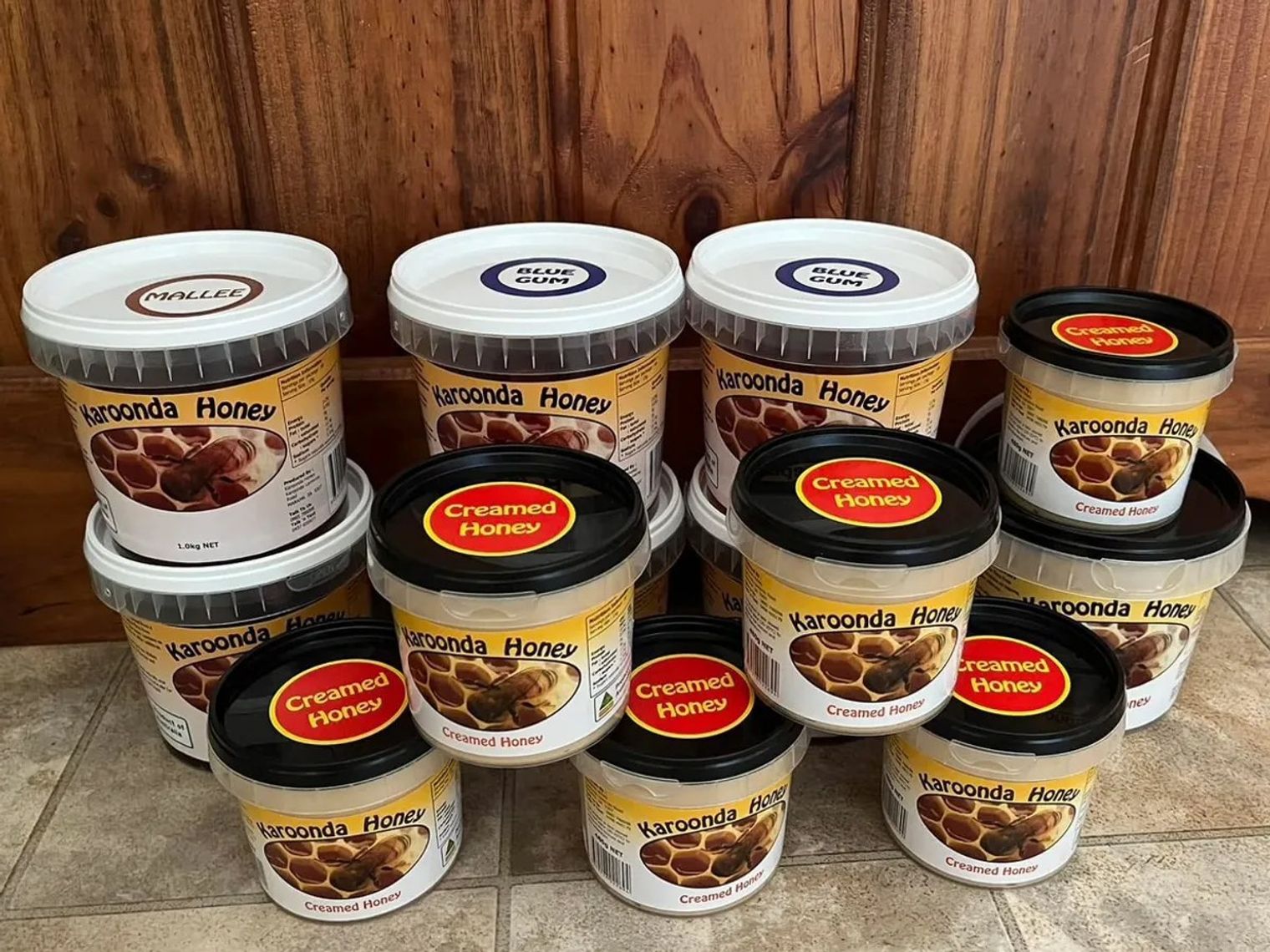 A stack of Karoonda Honey tubs on a tiled floor against a timber wall, labelled Mallee, Blue Gum and Creamed Honey