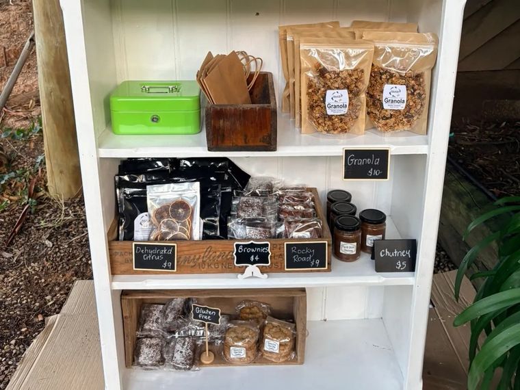 A white roadside cabinet at Palate Sensations in Greenhill, stocked with bagged granola, brownies, gluten free cookies and jars, with chalkboard price tags