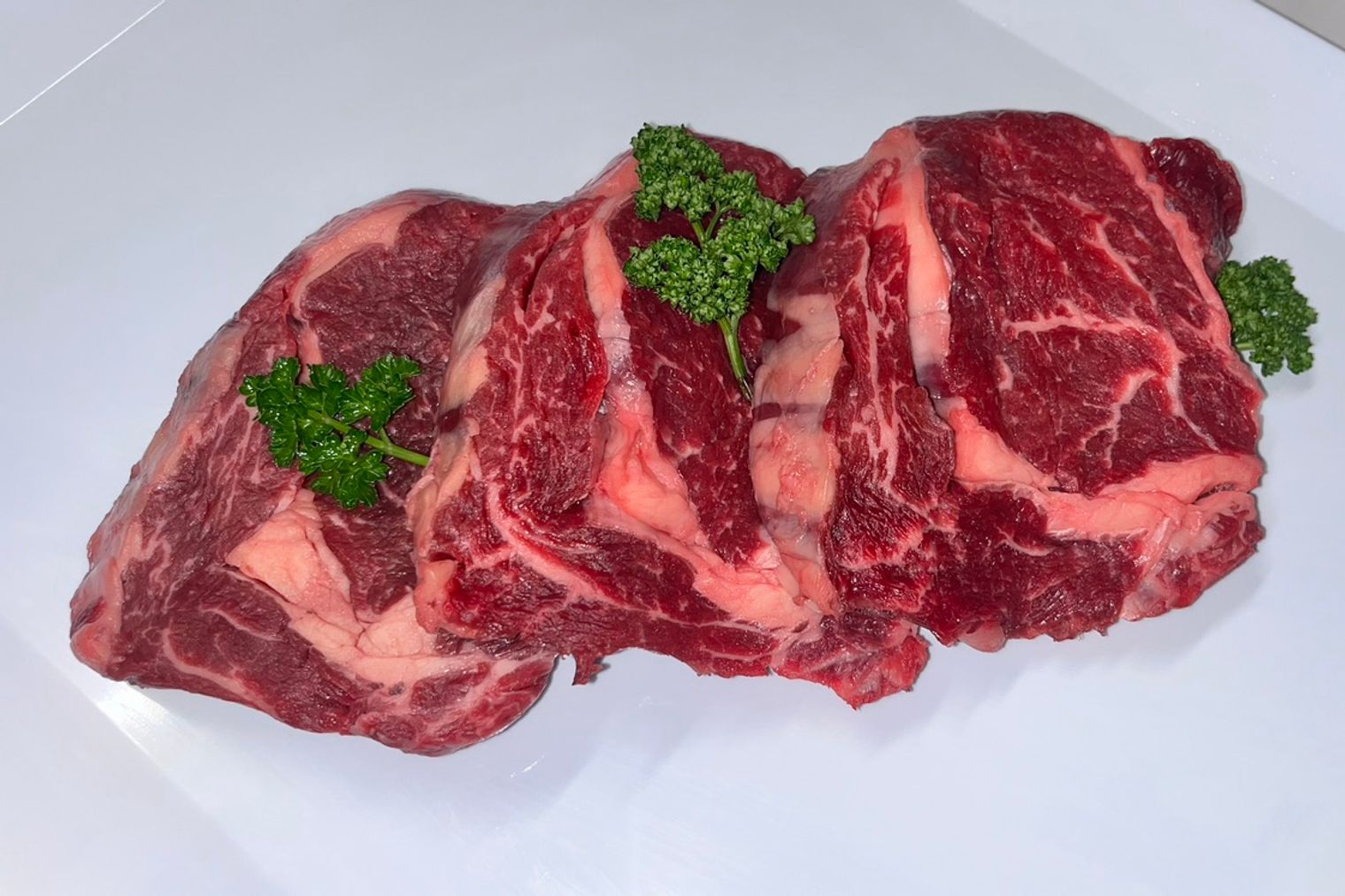 Three thick-cut marbled beef steaks with curly parsley on a white bench