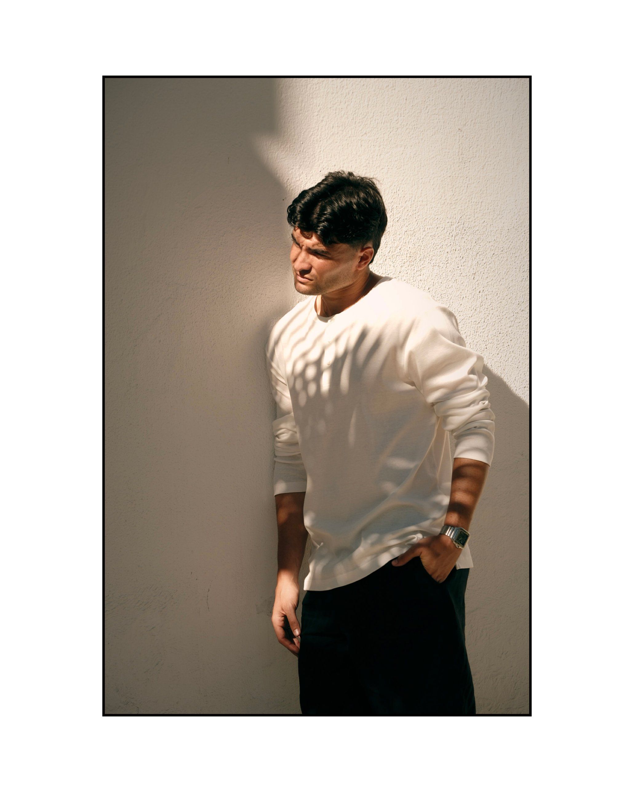 Aritz Aldasoro apoyado en una pared blanca con sombras de luz natural sobre la camiseta.