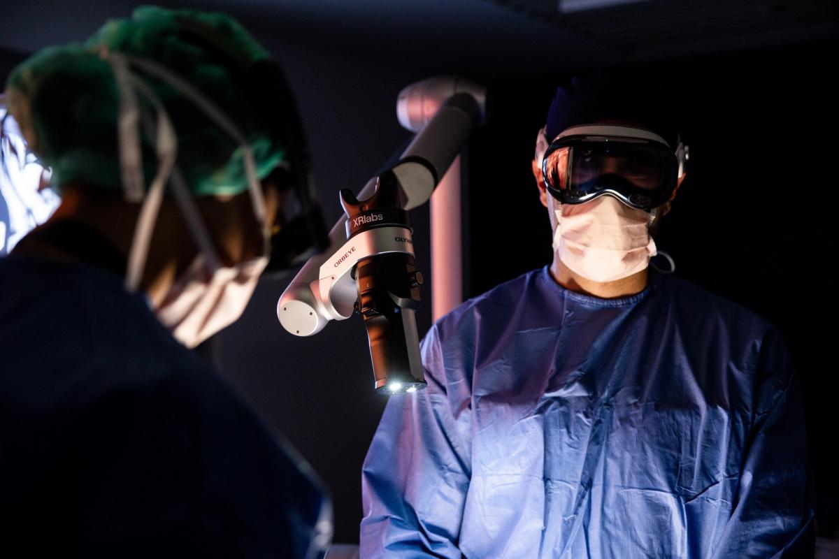 A surgeon wearing a head-mounted display stands beside an ORBEYE 4K 3D exoscope on an articulated arm in a darkened operating theatre, a colleague in scrubs opposite.