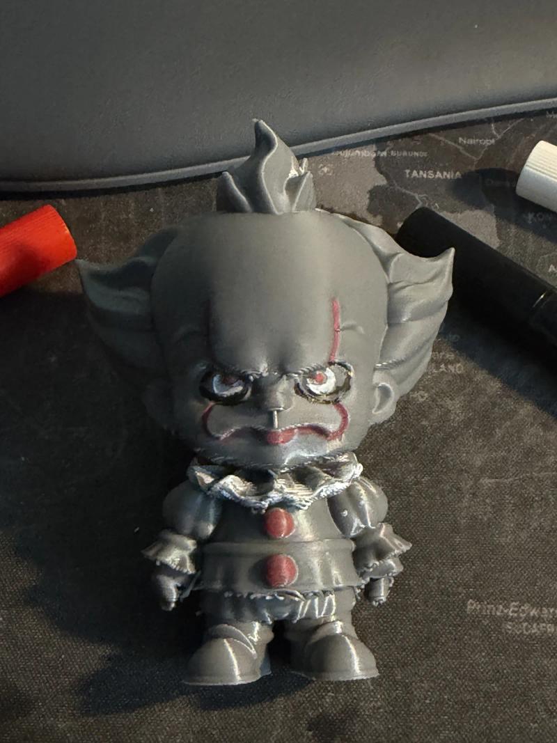 Chibi-Figur eines Clowns aus grauem PLA, direkt mit Acryl-Stiften bemalt – rote Linien im Gesicht, rote Knöpfe am Kostüm; daneben liegen die verwendeten Stifte
