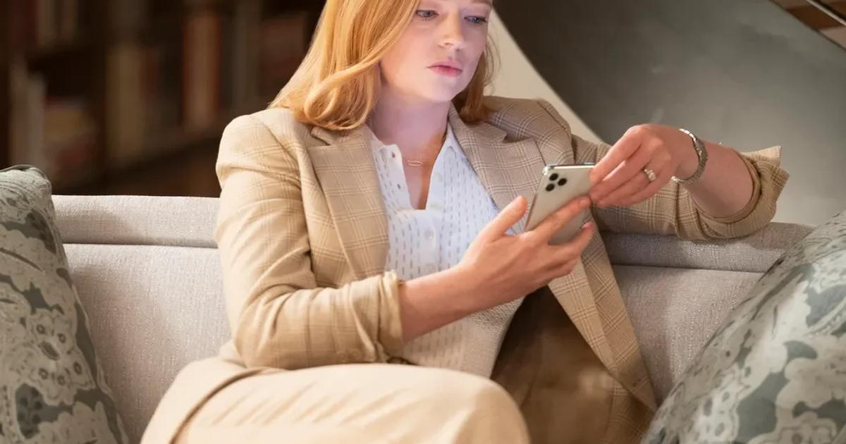 A woman in a cream trouser suit sitting on a pale sofa in a book lined room, looking down at the phone in her hands.