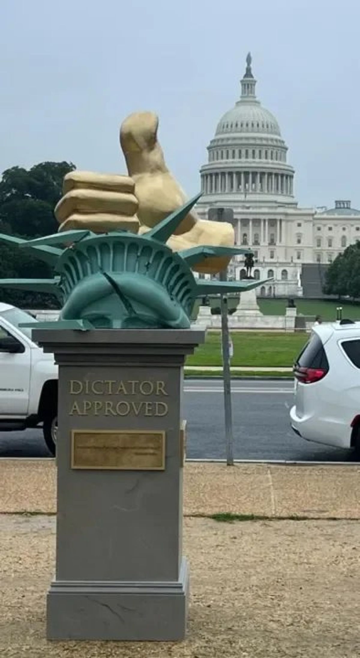 A sculpture of the Statue of Liberty inverted on a grey plinth with a plaque reading Dictator Approved, the United States Capitol dome behind it.