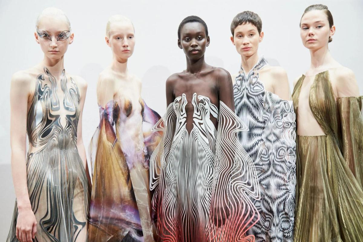 Five models standing in a row in sculptural dresses printed and pleated in silver, cream and blush, each shaped to flow around the body.
