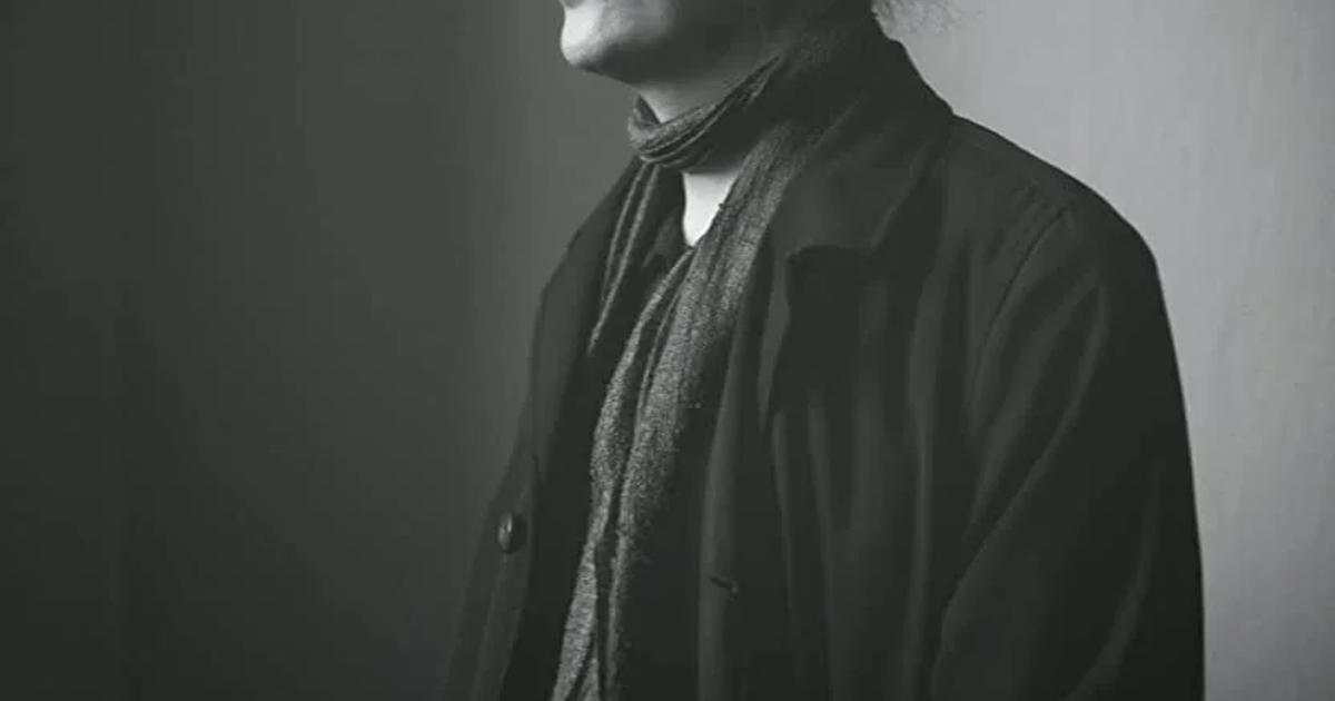 A black and white portrait of a man in a dark coat with his hair tied back, turned in profile and lit from one side against a plain grey background.