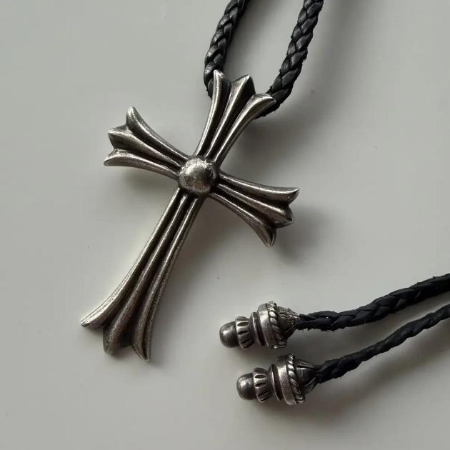 A heavy silver cross pendant with flared fleur tipped arms and a domed centre, strung on a braided black leather cord with silver end caps, on a pale grey surface.