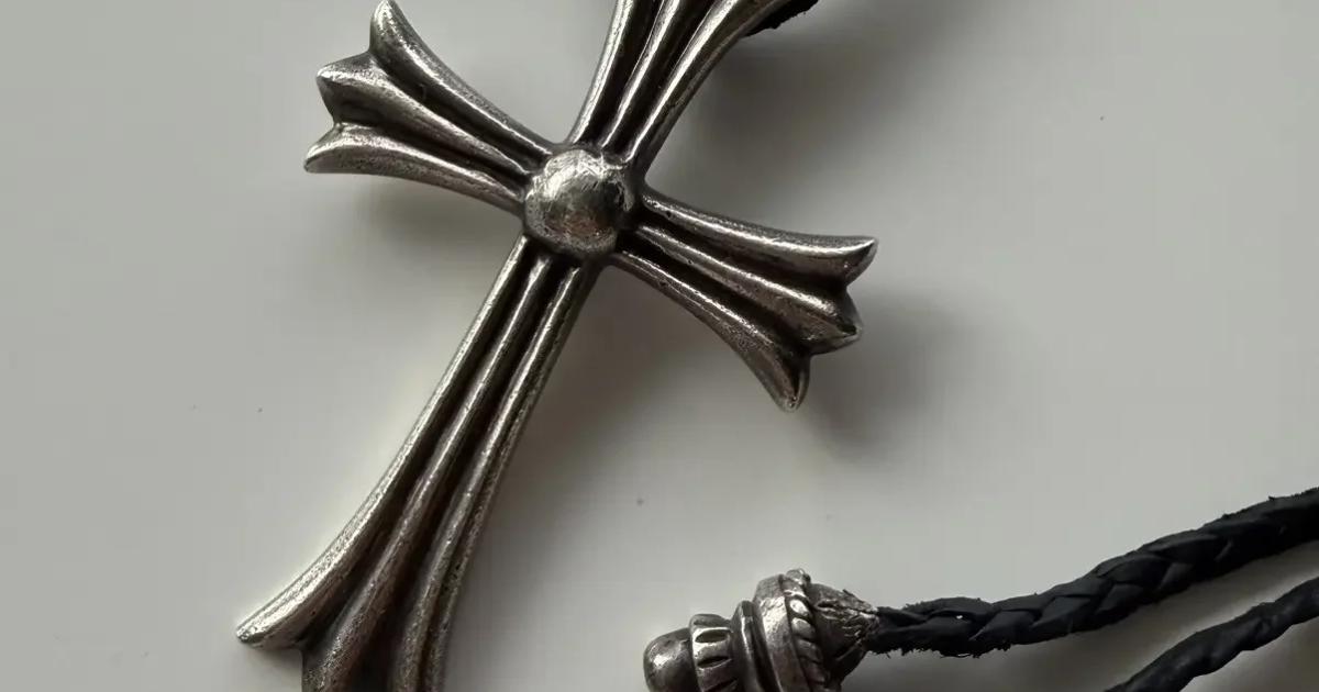 A heavy silver cross pendant with flared fleur tipped arms and a domed centre, strung on a braided black leather cord with silver end caps, on a pale grey surface.