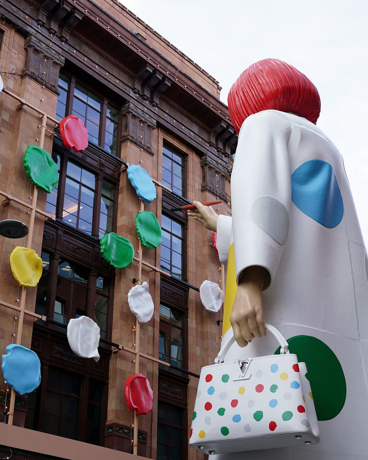 A giant figure of Kusama in a red wig and white dotted coat mounted on the outside of a department store, holding a branded handbag and appearing to paint dots on the building.