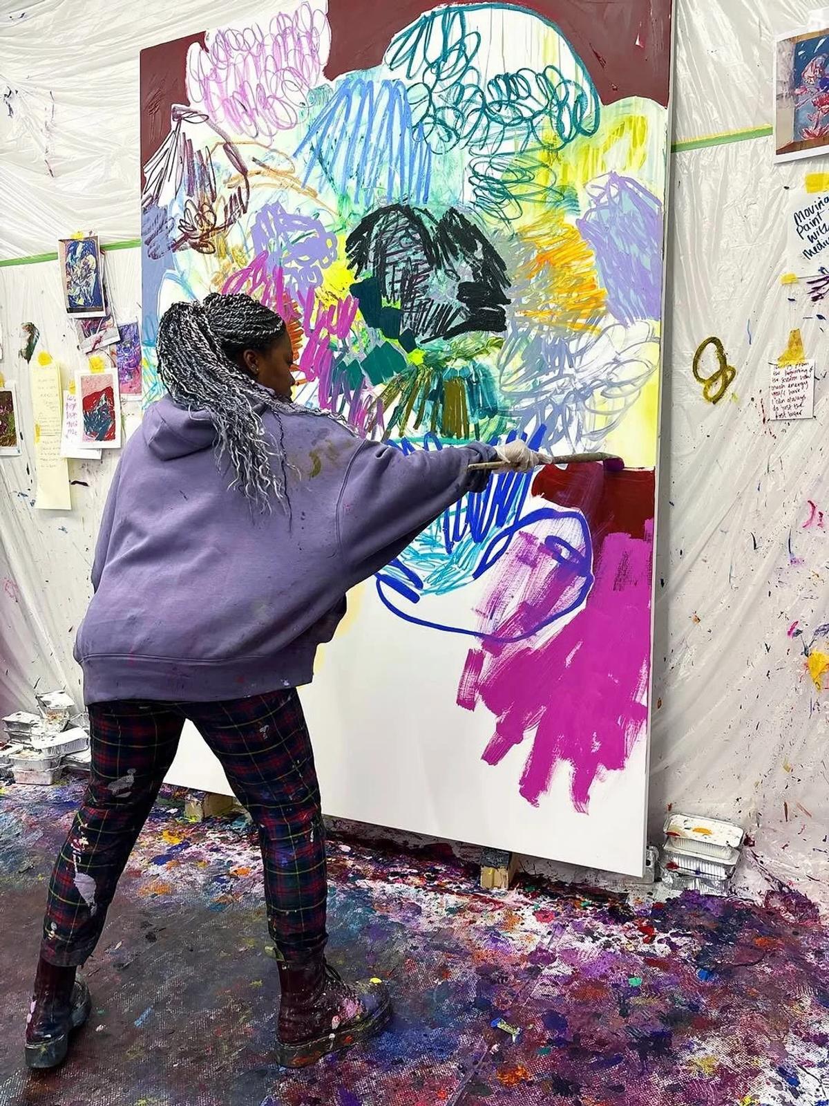 An artist in a purple hoodie working on a large canvas in a studio, laying broad pink and blue strokes over looping drawn lines, paint spattered across the floor.