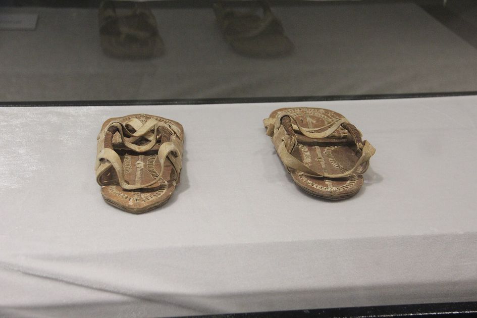 A pair of worn leather sandals with ornamented straps, displayed on a white cloth inside a museum vitrine.