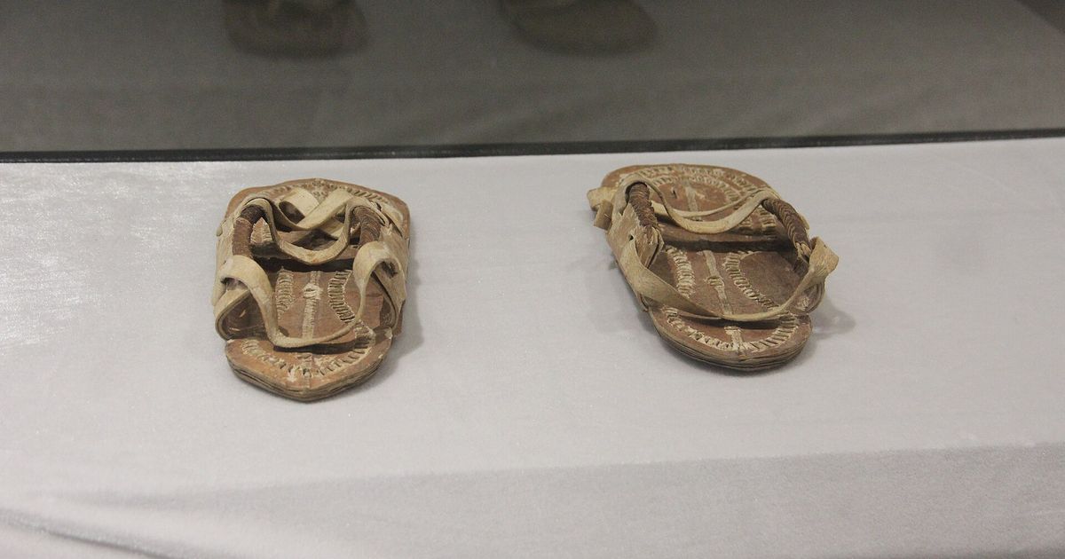 A pair of worn leather sandals with ornamented straps, displayed on a white cloth inside a museum vitrine.