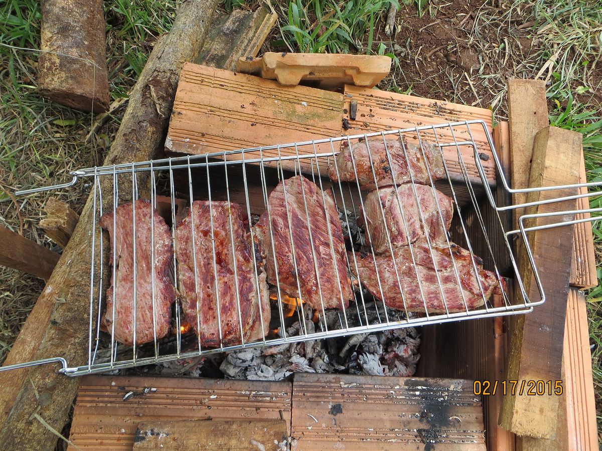 A wire grill basket of beef held over glowing embers in a fire pit improvised from roof tiles and split firewood on grass. No people are in the frame.