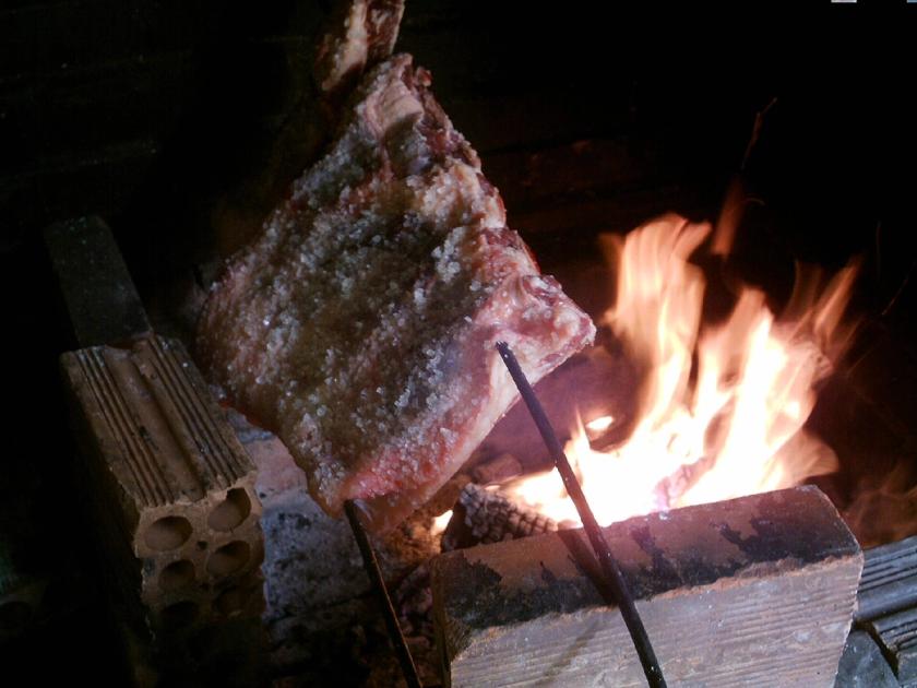 A salt-crusted beef rib on an iron skewer angled over an open wood fire built between loose bricks, flames to one side. No people are in the frame.