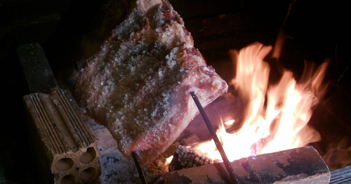 A salt-crusted beef rib on an iron skewer angled over an open wood fire built between loose bricks, flames to one side. No people are in the frame.