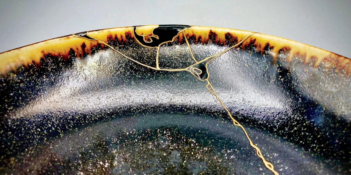 A close view of a dark ceramic bowl rim repaired with fine gold seams running through the glaze, the surface speckled cream and amber.