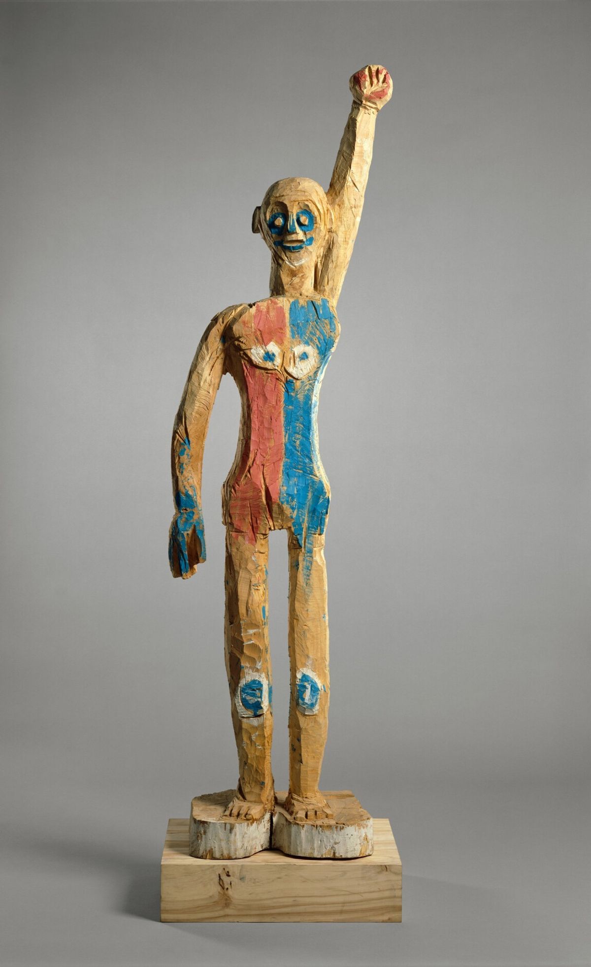 An eight-foot carved limewood figure by Georg Baselitz, roughly hewn, painted in patches of red, blue and white, standing on a wooden block with its left arm raised straight up.