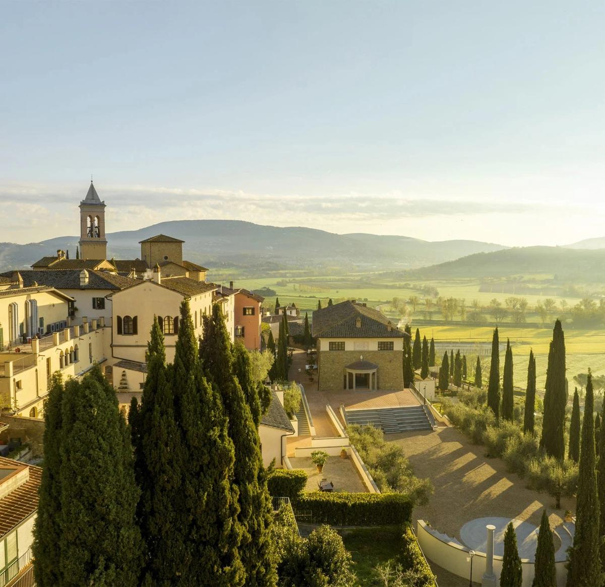 An Italian hill town of pale stone buildings and cypress trees seen from above at dawn, farmland and hills stretching into the distance.